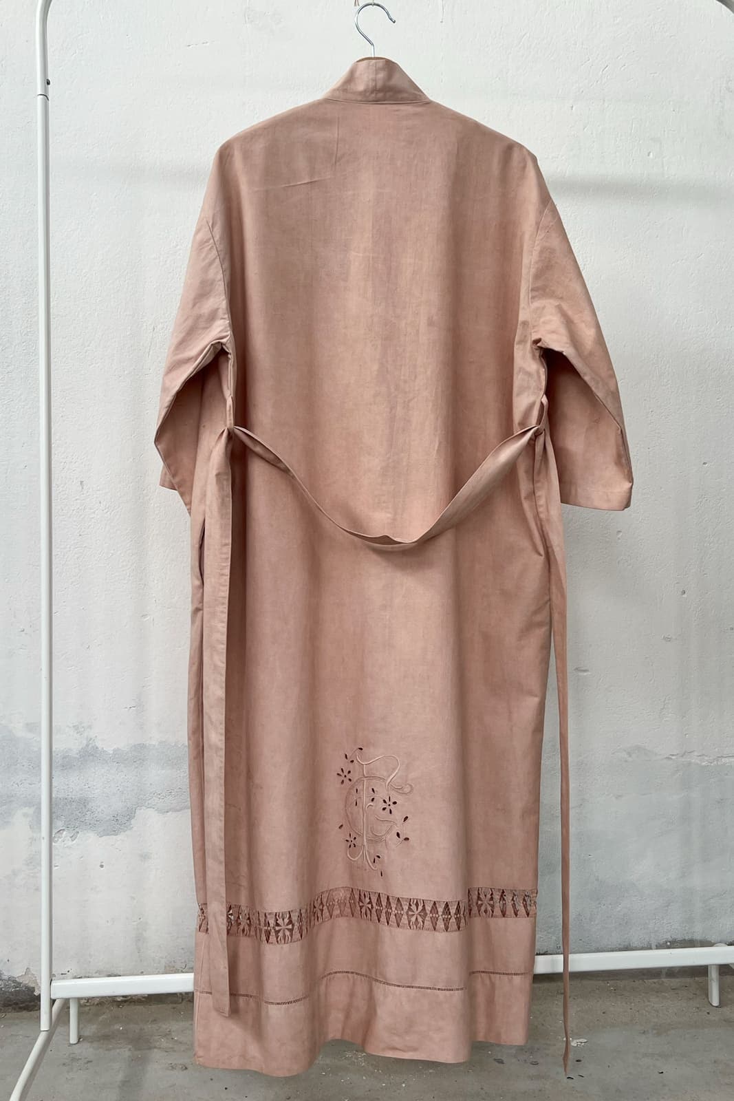 Long antique linen kimono naturally dyed in soft pink, hanging on a clothing rack, with a wooden hanger and white wall in the background. 