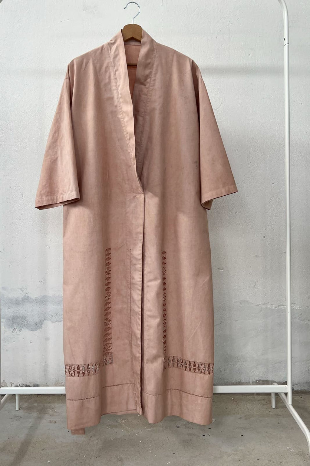Long antique linen kimono dress naturally dyed in soft pink, hanging on a clothing rack, with a wooden hanger and white wall in the background. 