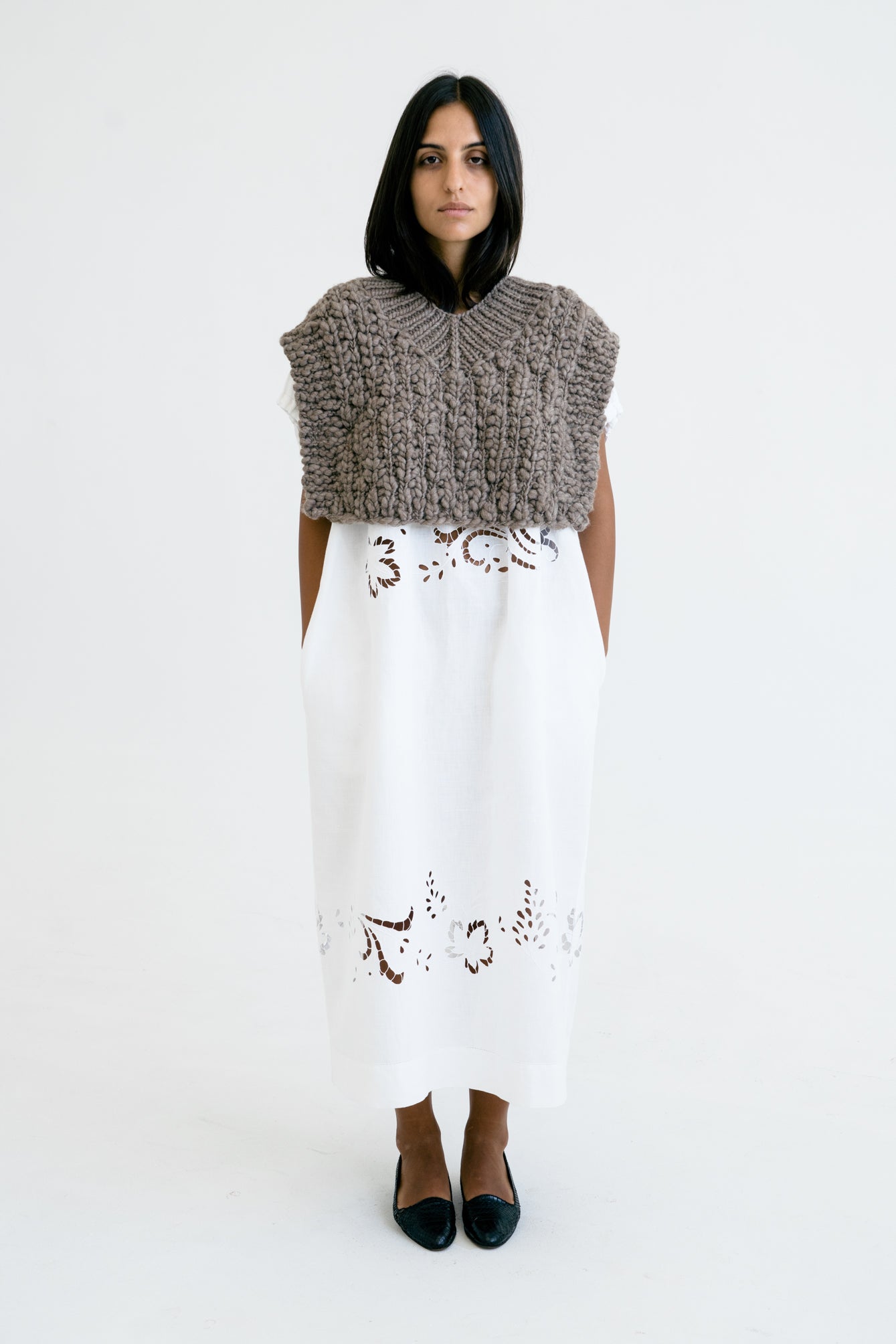 Woman wearing a knitted sleeveless top and white dress with lace details on a white background