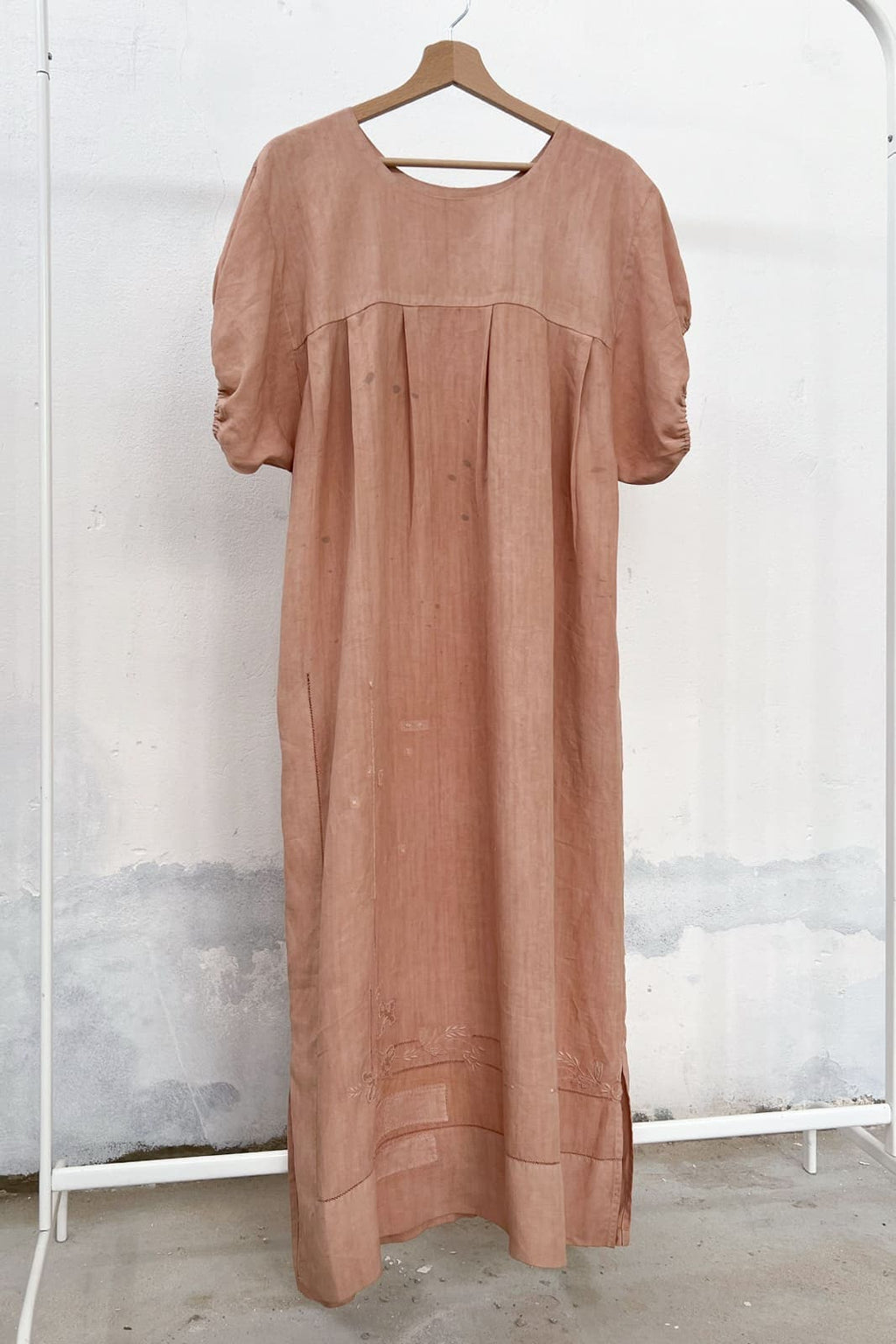 Long antique linen dress naturally dyed in soft pink, with avocado, hanging on a clothing rack, with a wooden hanger and white wall in the background. 