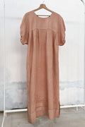 Long antique linen dress naturally dyed in soft pink, with avocado, hanging on a clothing rack, with a wooden hanger and white wall in the background. 