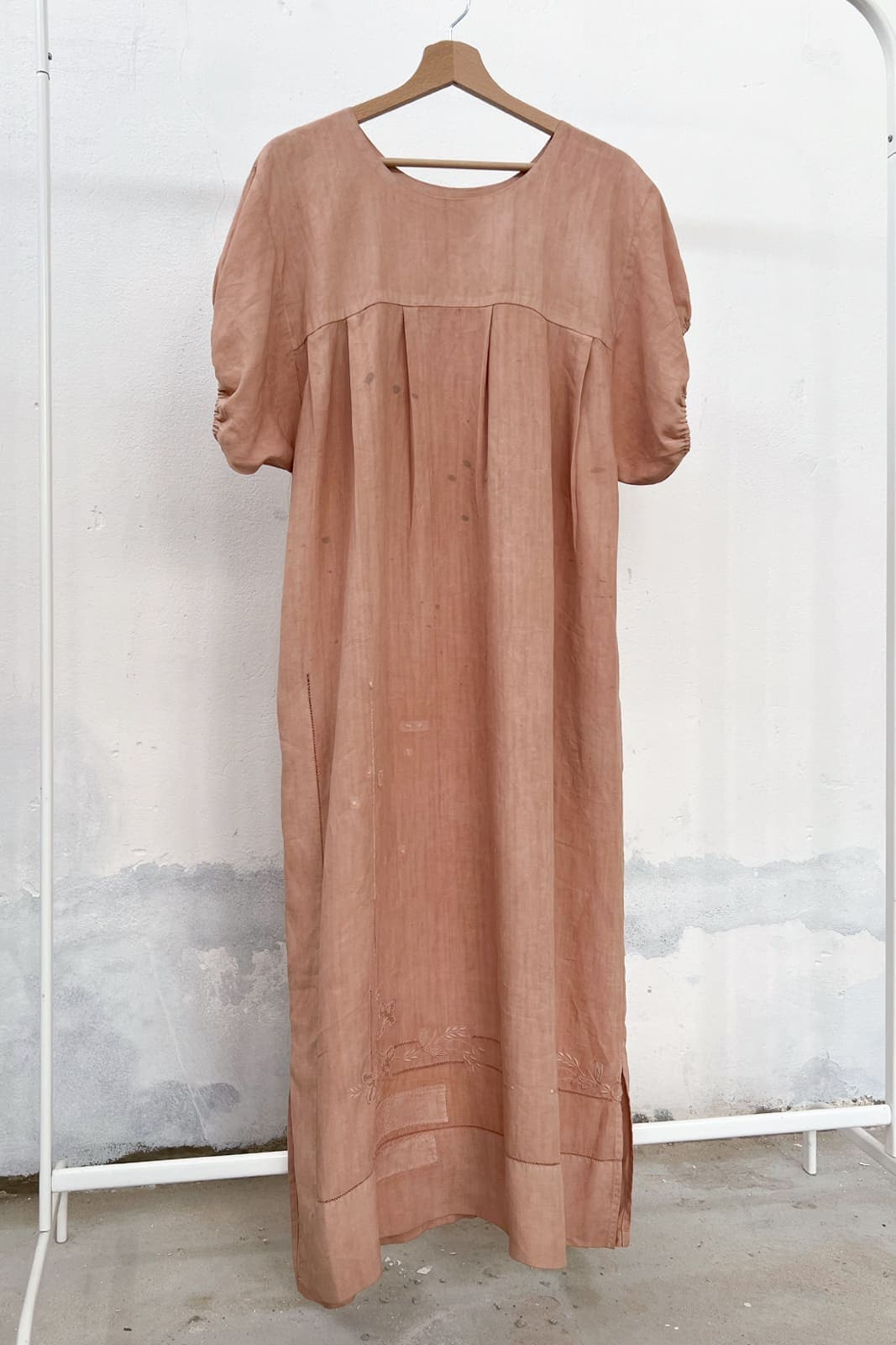Long antique linen dress naturally dyed in soft pink, with avocado, hanging on a clothing rack, with a wooden hanger and white wall in the background. 