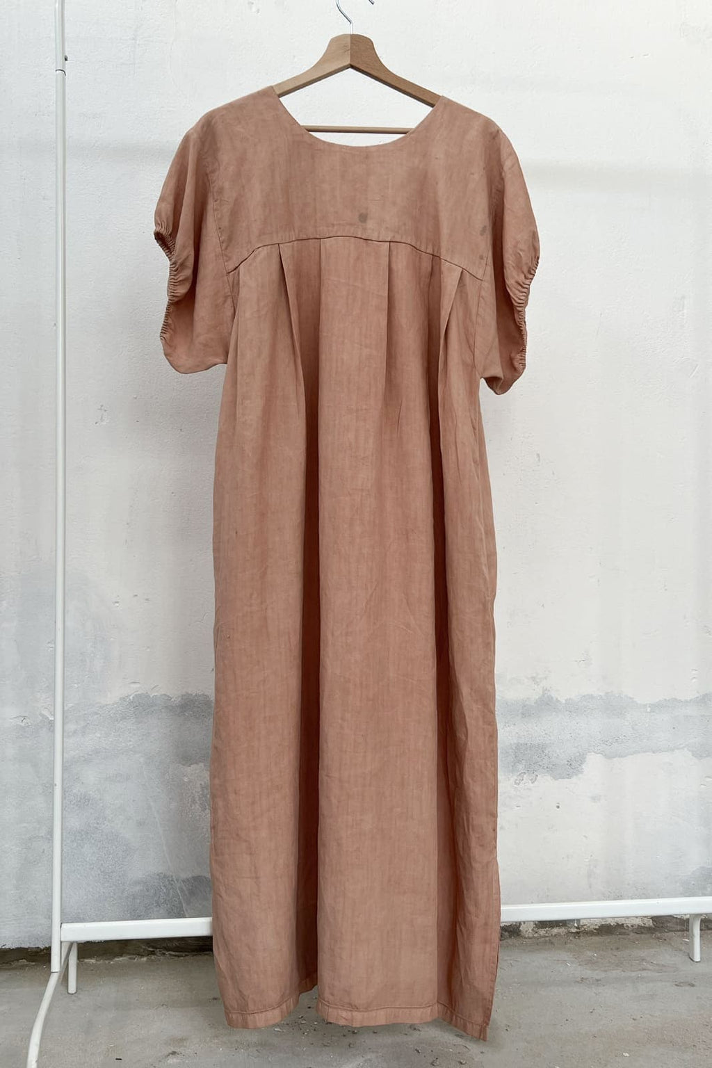 Long antique linen dress naturally dyed in soft pink, with avocado, hanging on a clothing rack, with a wooden hanger and white wall in the background. 