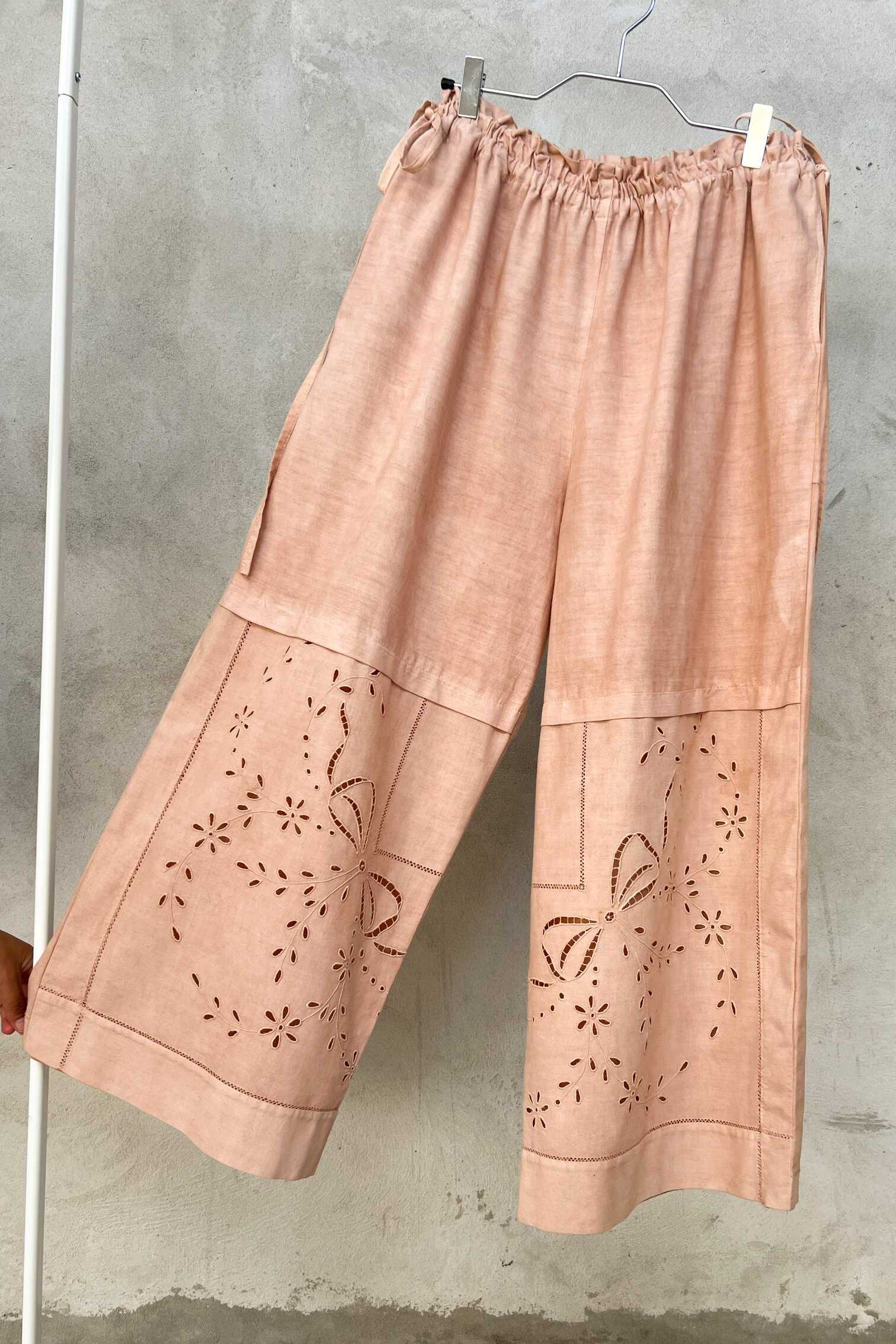 Wide-leg dusty pink linen trousers hanging on a metal hanger against a textured grey wall, featuring floral cutwork embroidery on the lower legs and an adjustable gathered waist.