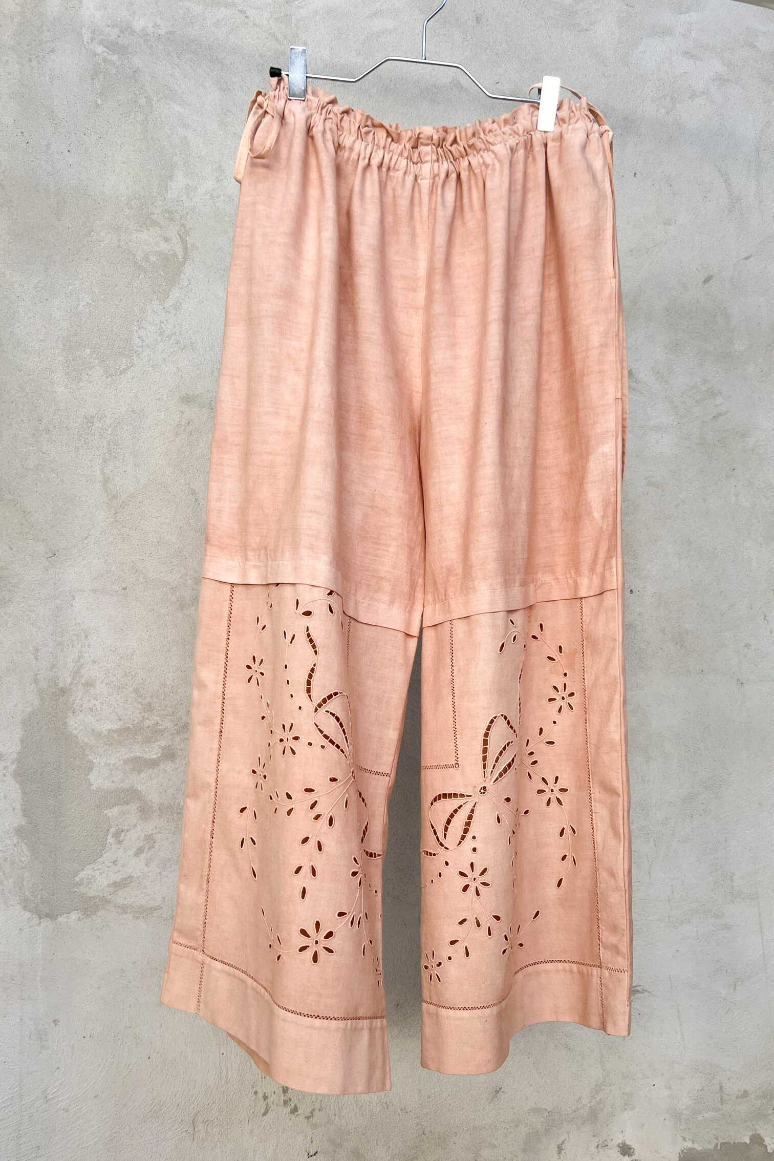 Wide-leg dusty pink linen trousers hanging on a metal hanger against a textured grey wall, featuring 