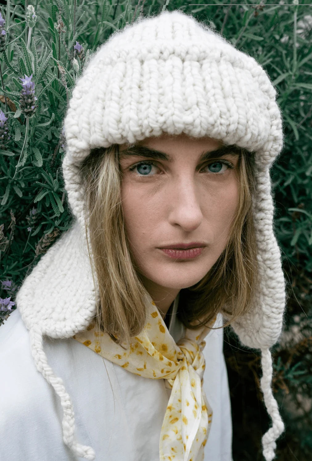 Person wearing a white knitted hat with earflaps against a blurred natural background